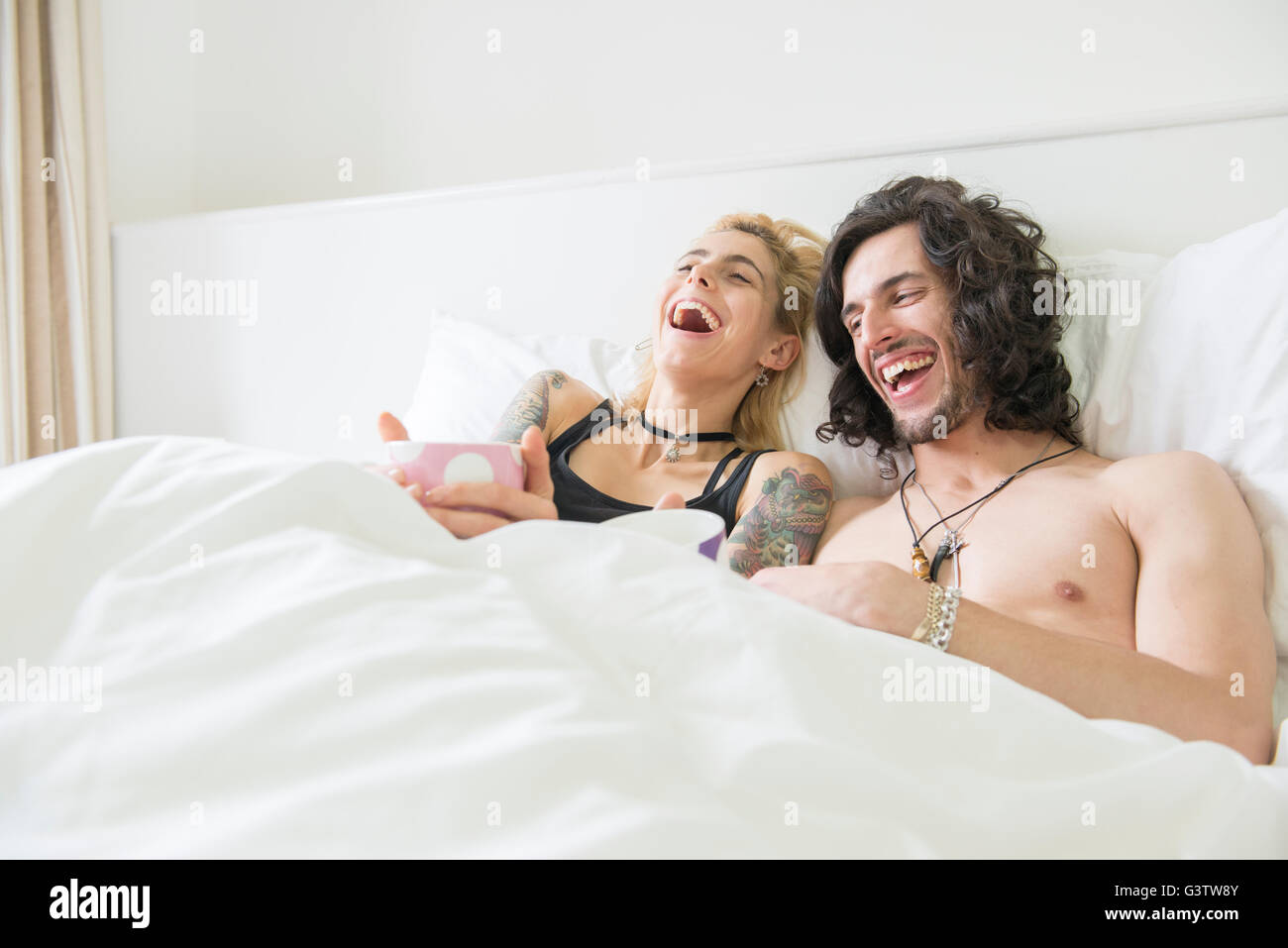 A cool young tattooed couple lying in a bed enjoying a cup of tea and laughing. Stock Photo