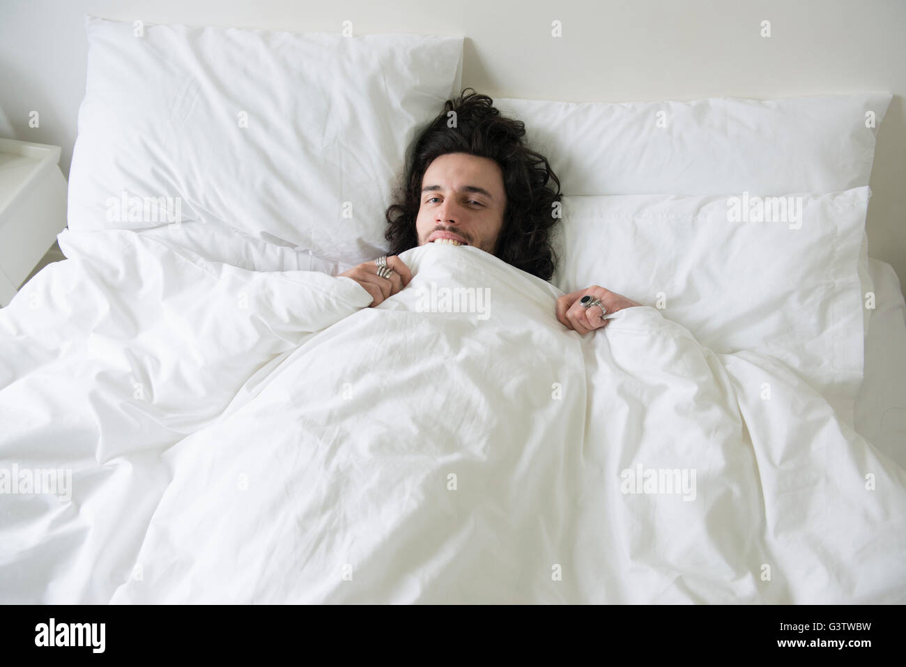 A cool young man lying in a bed playing peekaboo with the covers to the camera. Stock Photo