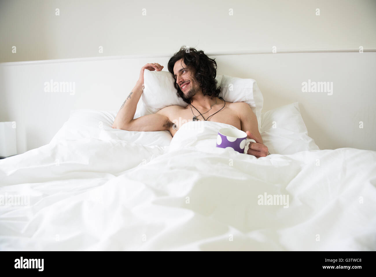 A young man with long hair lying in a bed with a cup of tea looking happy. Stock Photo