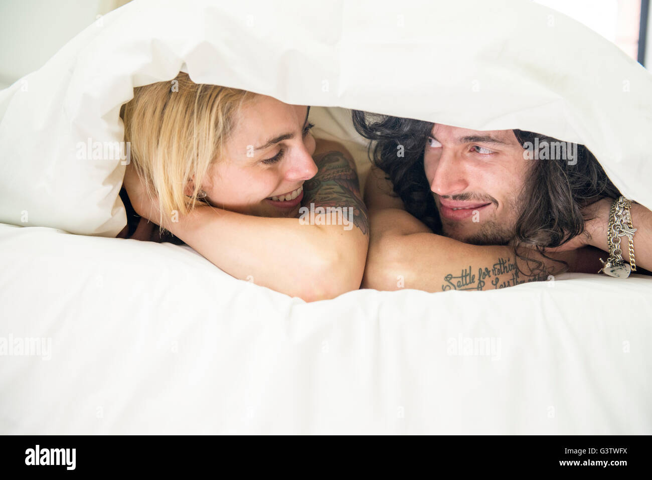 A cool young tattooed couple peeking out from under a duvet on a bed. Stock Photo