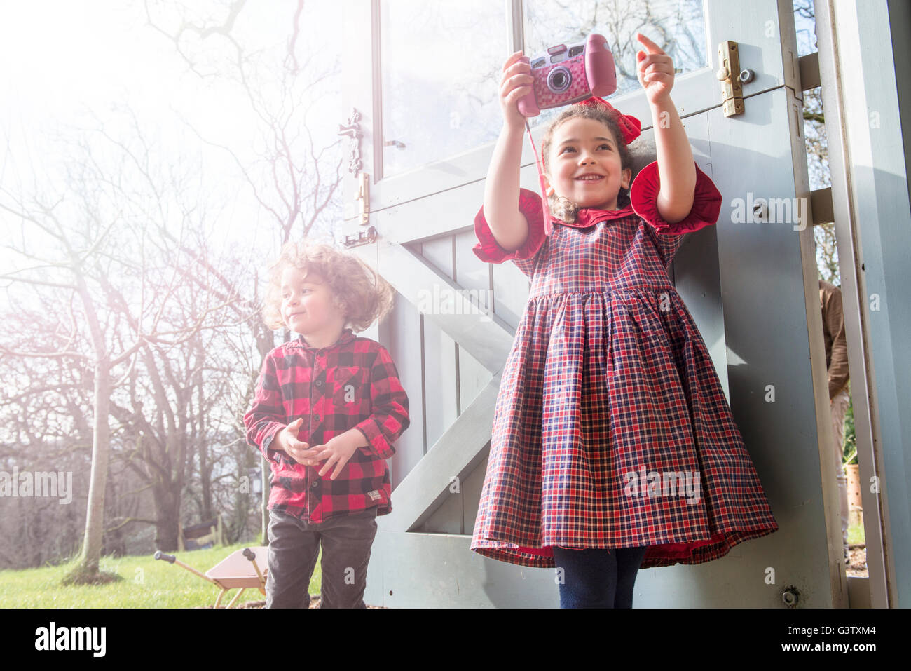 A six year old girl standing with her brother at a back door taking pictures with a toy camera. Stock Photo