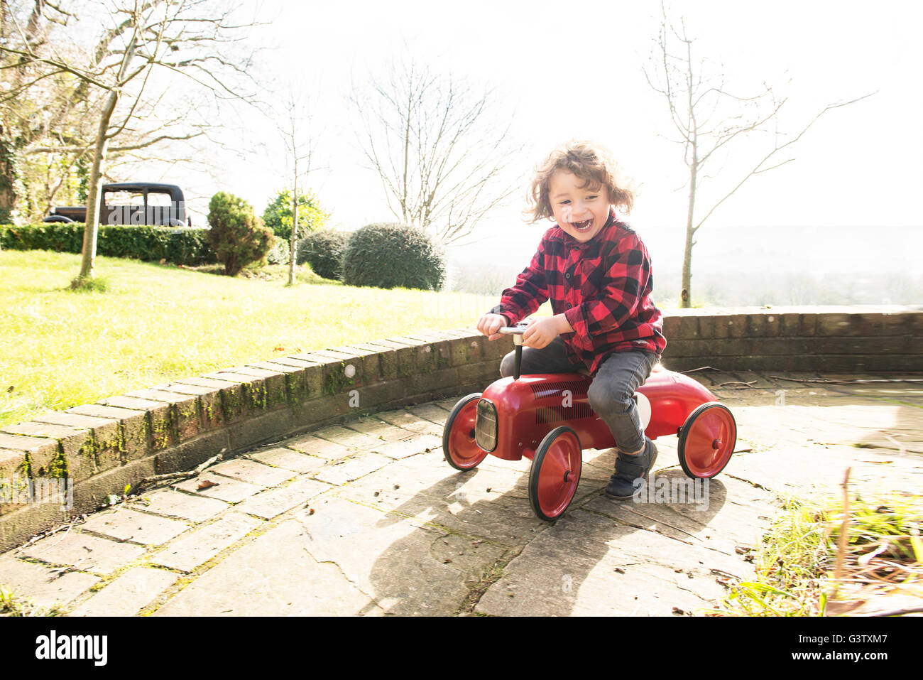 A four year old boy riding a toy tractor around the garden in a check shirt. Stock Photo