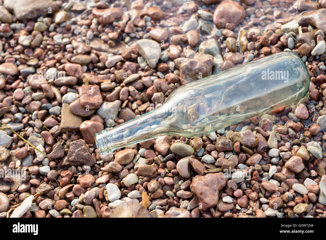 Single empty clear glass bottle on stones at the edge of the waterside. Stock Photo