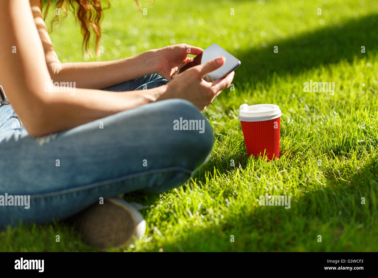 Young woman with red disposable paper cup of coffee using tablet pc on a green lawn in a park Stock Photo