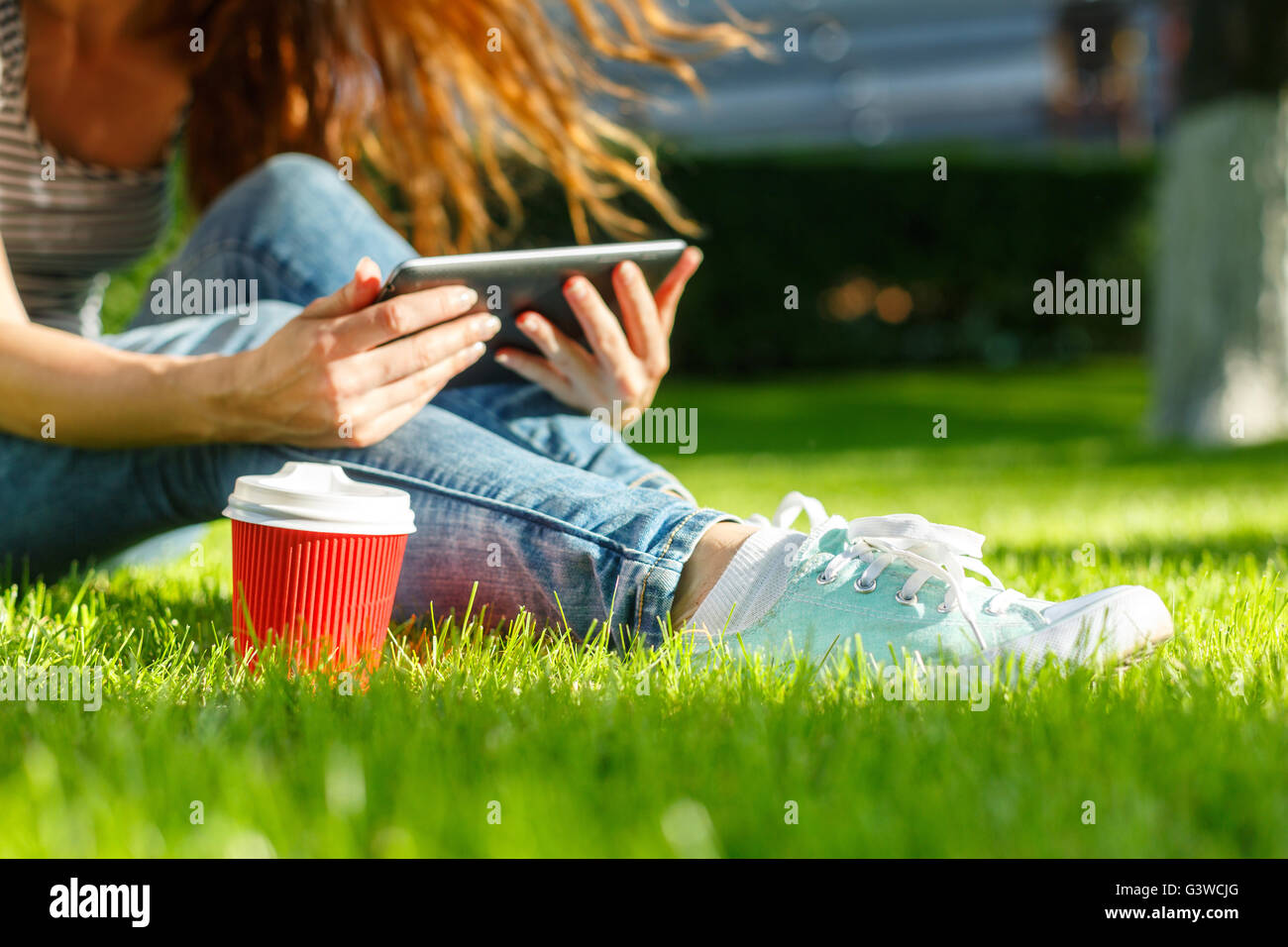Young woman with red disposable paper cup of coffee using tablet pc on a green lawn in a park Stock Photo
