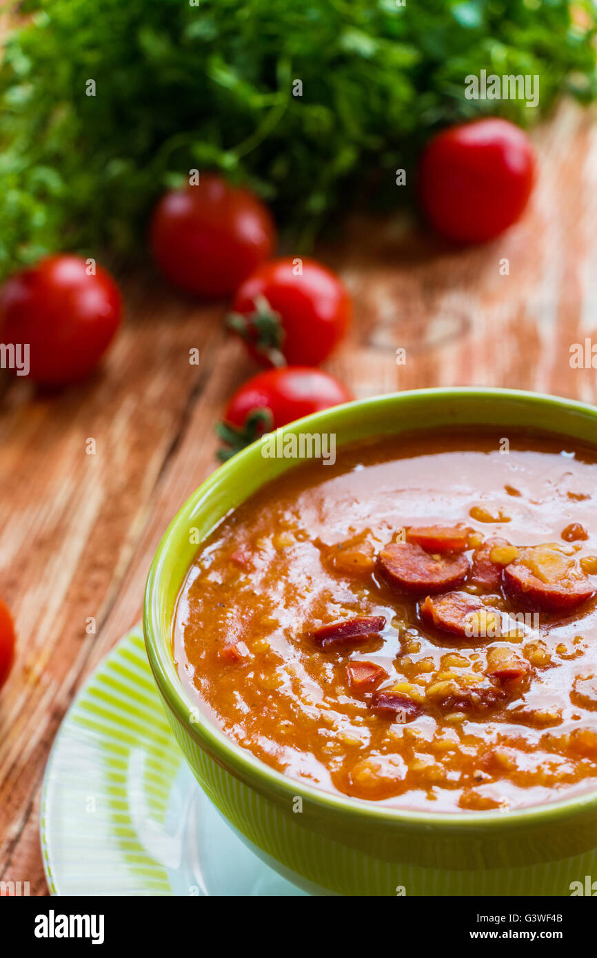 Soup: tomato soup with smoked sausage, tomatoes and lentil. Green bowl on wooden table. Small cherry tomatoes and green lettuce Stock Photo