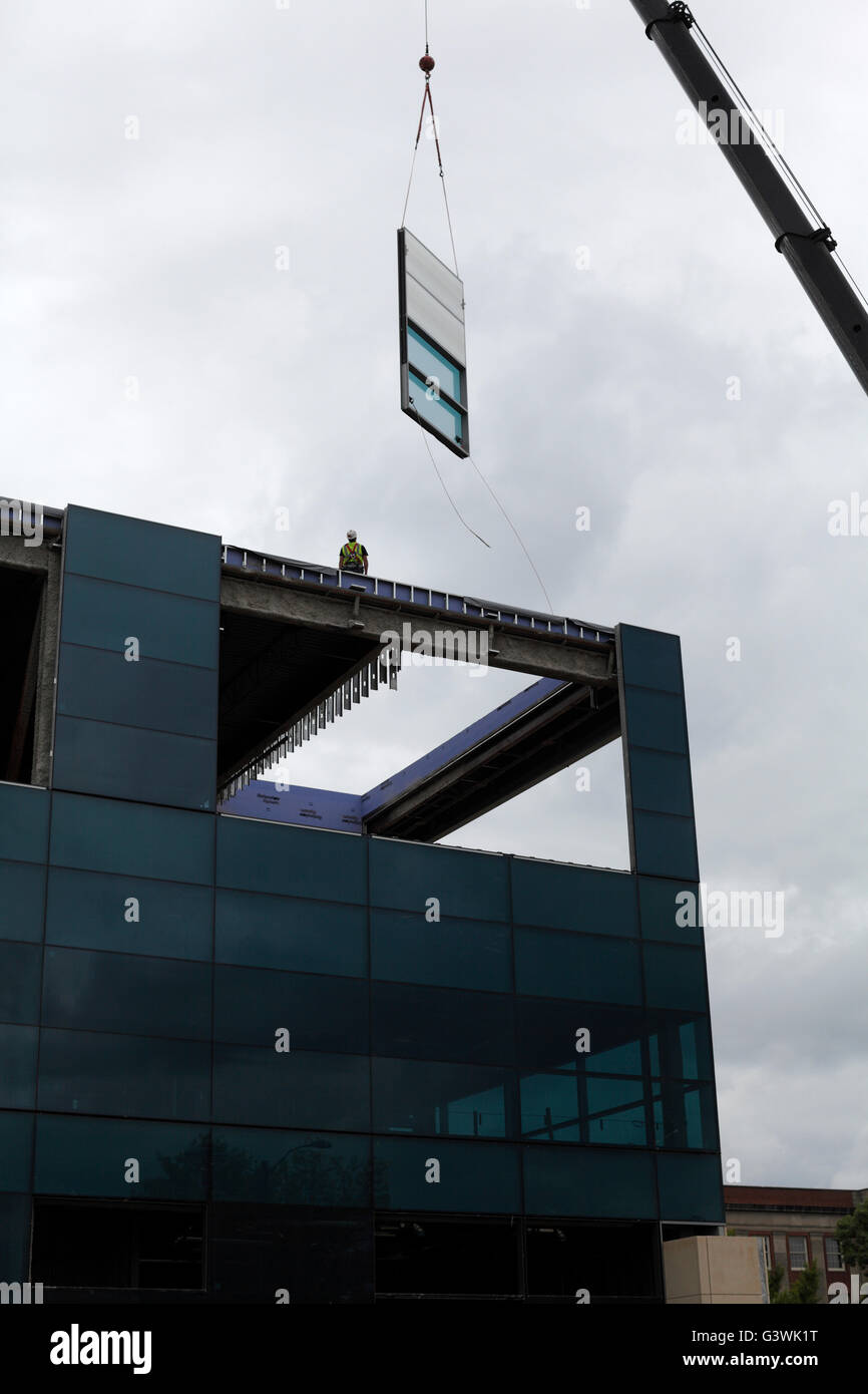 A person stands atop a building while a crane lowers a prefabricated window panel into place. Stock Photo