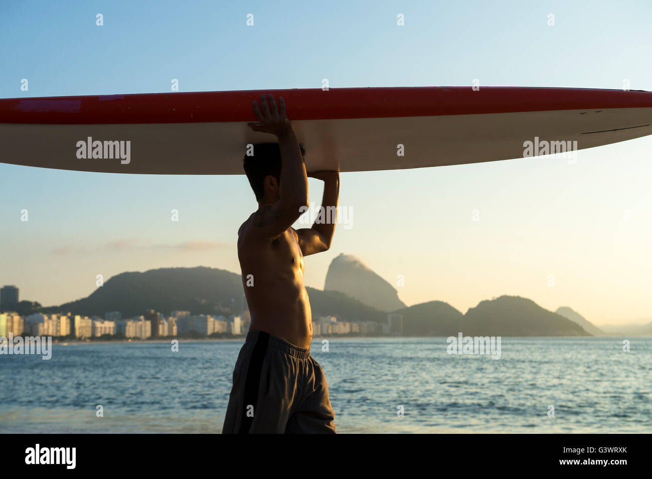 RIO DE JANEIRO - APRIL 5, 2016: A young Brazilian man on Copacabana Beach balances a surfboard on his head at sunrise. Stock Photo