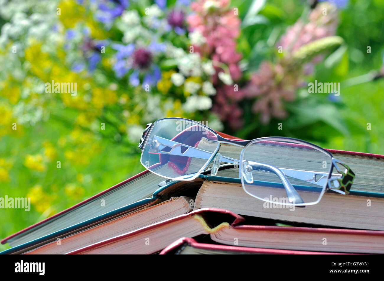 Reading glasses with a stack of inverted open books on the background of bouquet of wildflowers and green grass Stock Photo