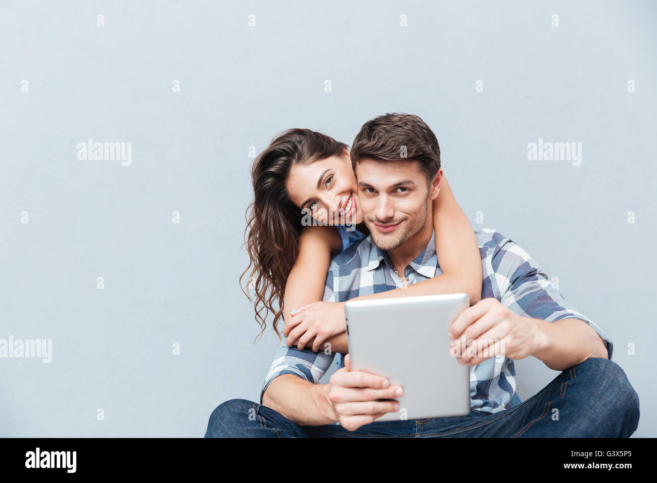 Happy couple sitting at home and using digital tablet isolated on gray ...