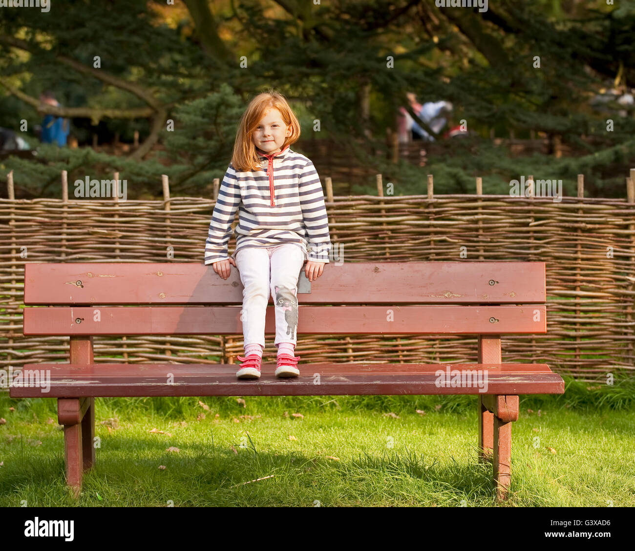 Sitting on a Bench. A young girl with red hair sits on the back of a park bench confronting the camera. Stock Photo