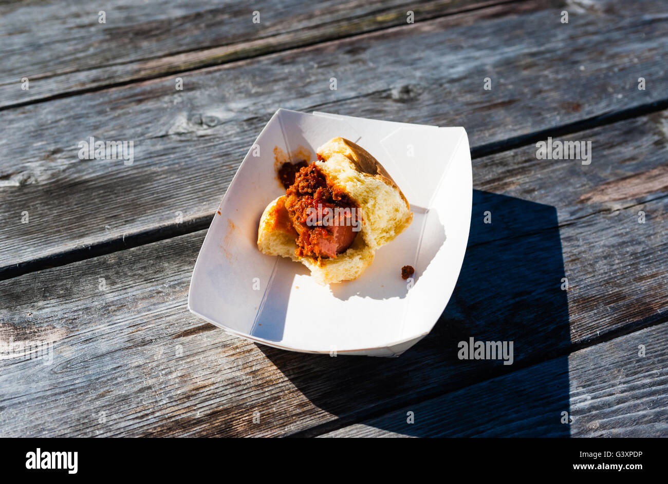 Remaining piece of eaten sausage and chili on a bun in a paper tray, on weathered gray picnic table. Stock Photo