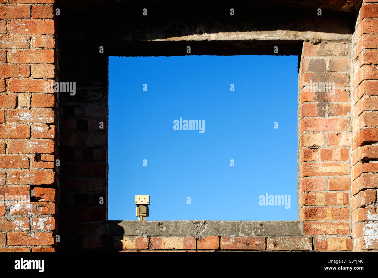 A Danbo Danboard fictional Robot Character framed against a blue sky in an empty window in an abandoned building Stock Photo