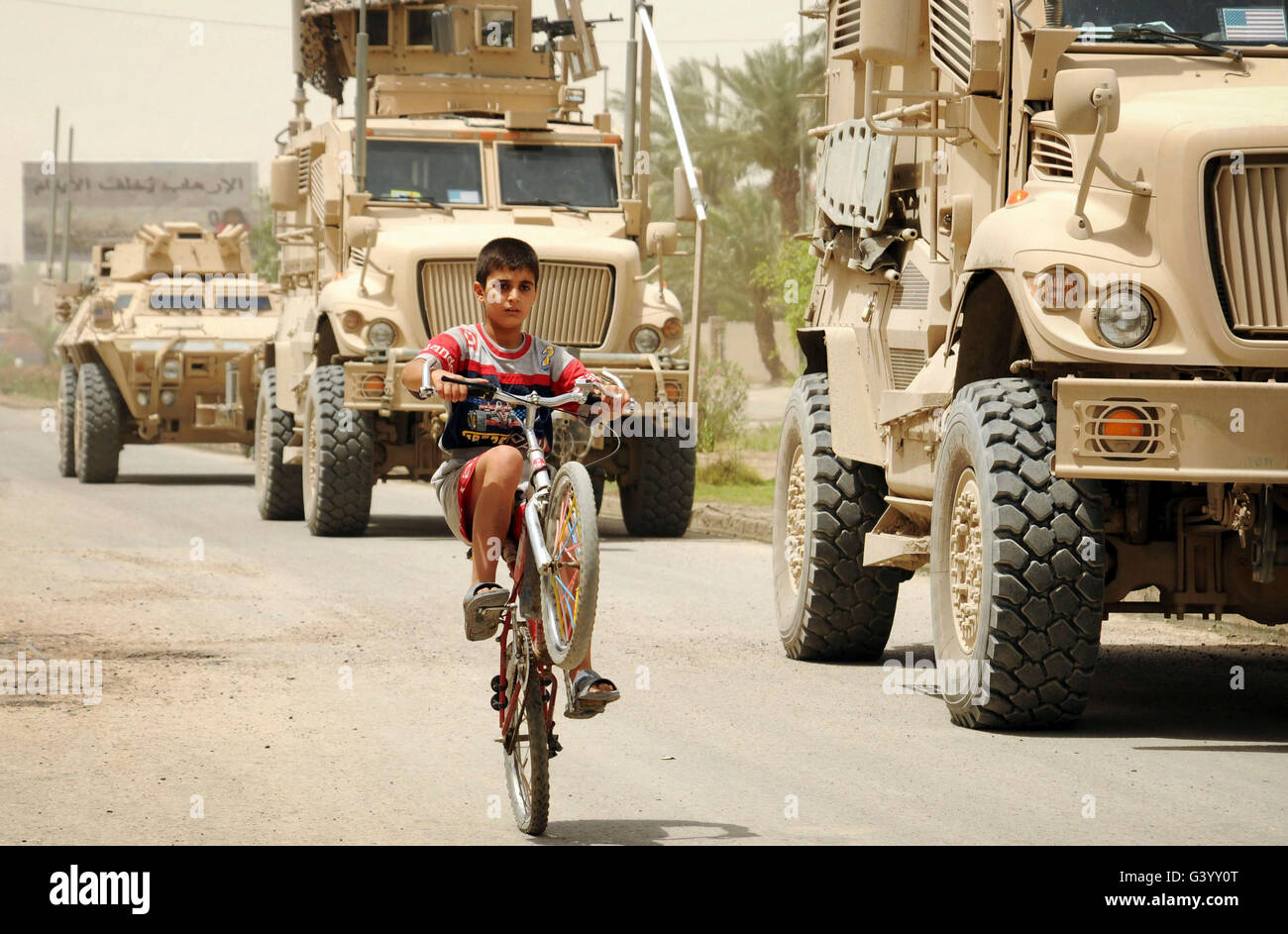 An Iraqi boy rides his bike past a U.S. Military vehicle patrol. Stock Photo
