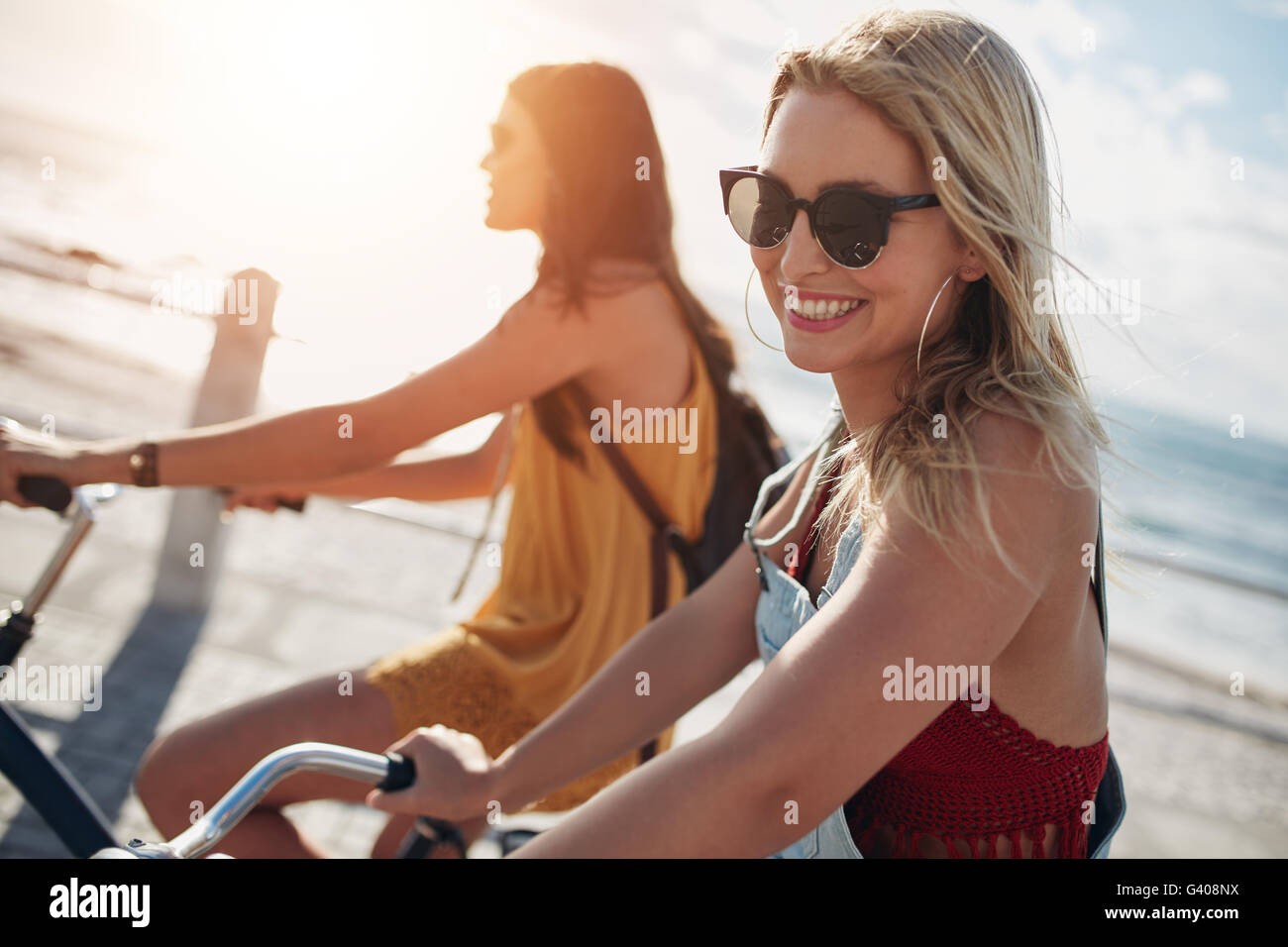 Smiling young woman riding bicycle with her friend on a sunny day. Two female friends cycling on a seaside promenade. Stock Photo