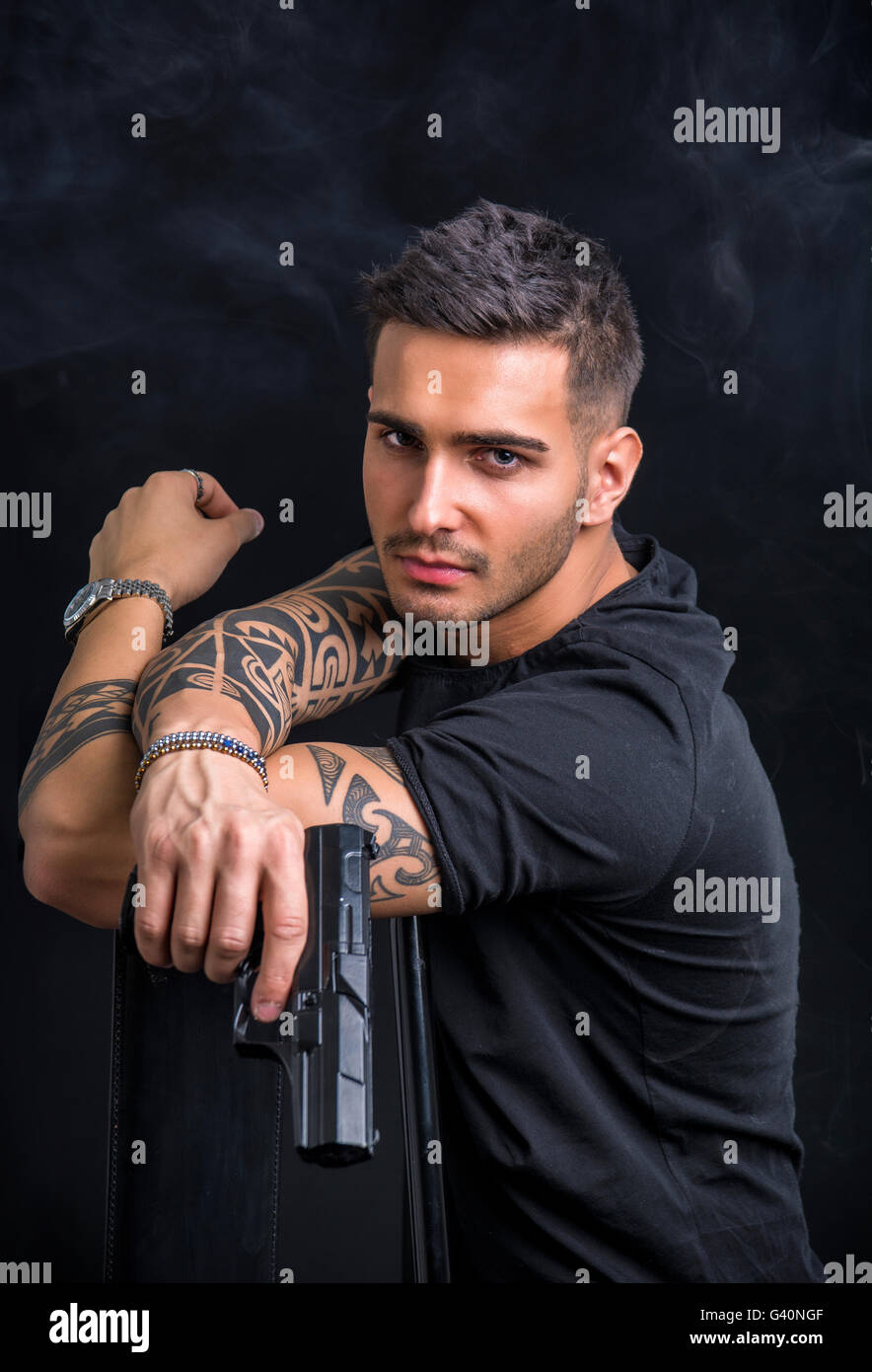Young handsome man holding a hand gun, wearing black t-shirt, on dark background in studio Stock Photo