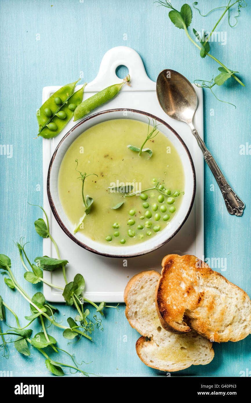 Light summer green pea cream soup in bowl with sprouts, bread toasts and spices. White ceramic board in the center, turquoise bl Stock Photo