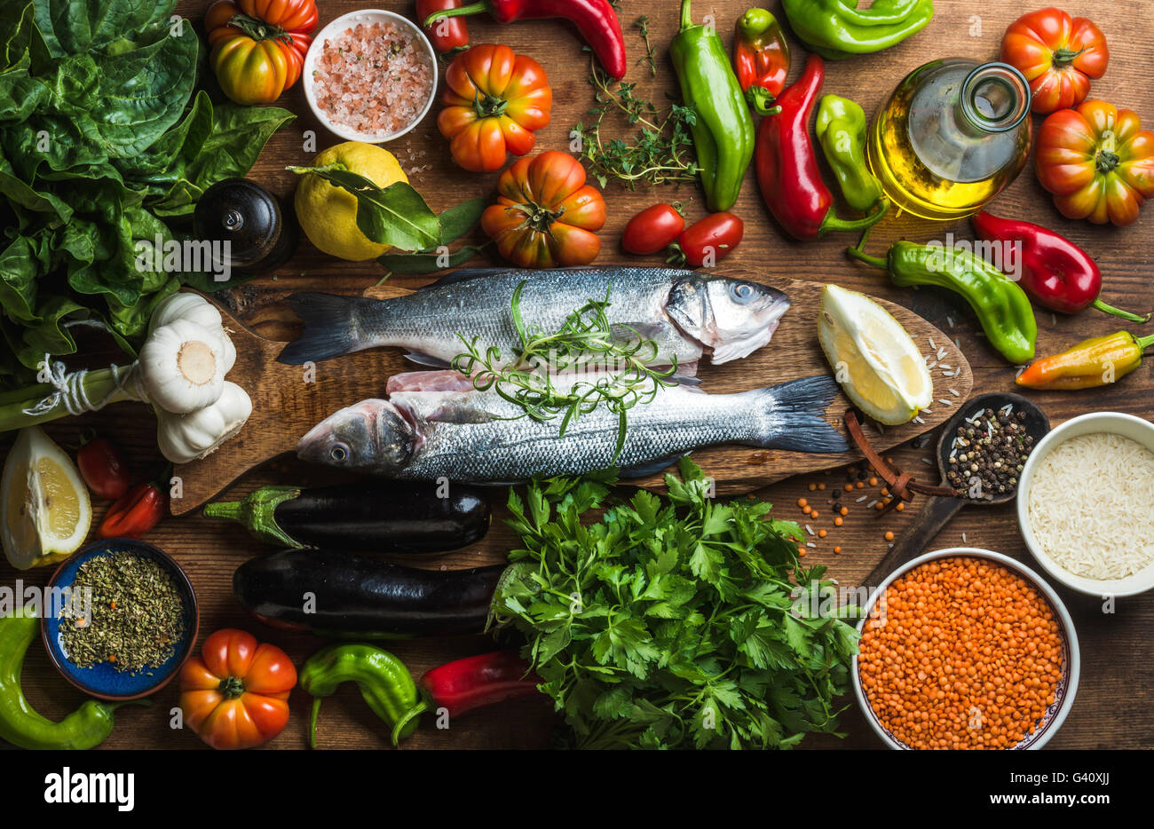 Raw uncooked seabass fish with vegetables, grains, herbs and spices on chopping board over rustic wooden background, top view Stock Photo