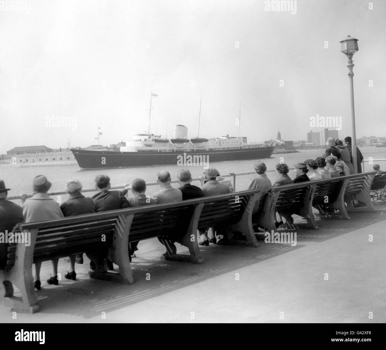 People sunning themselves on the seafront seats watch the dark blue Royal Yacht Britannia as she sails from Portsmouth on her way to London, where she will be boarded by Princess Margaret and Antony Armstrong-Jones after their wedding for a honeymoon cruise to the Caribbean. Stock Photo