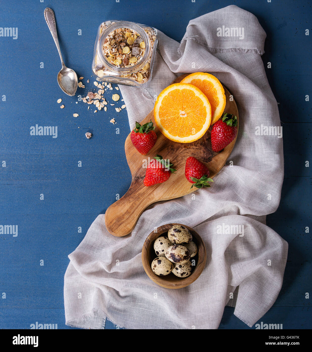 Ingredients for healthy breakfast. Quail eggs, jar of muesli, fresh orange and strawberries on olive wood chopping board with te Stock Photo