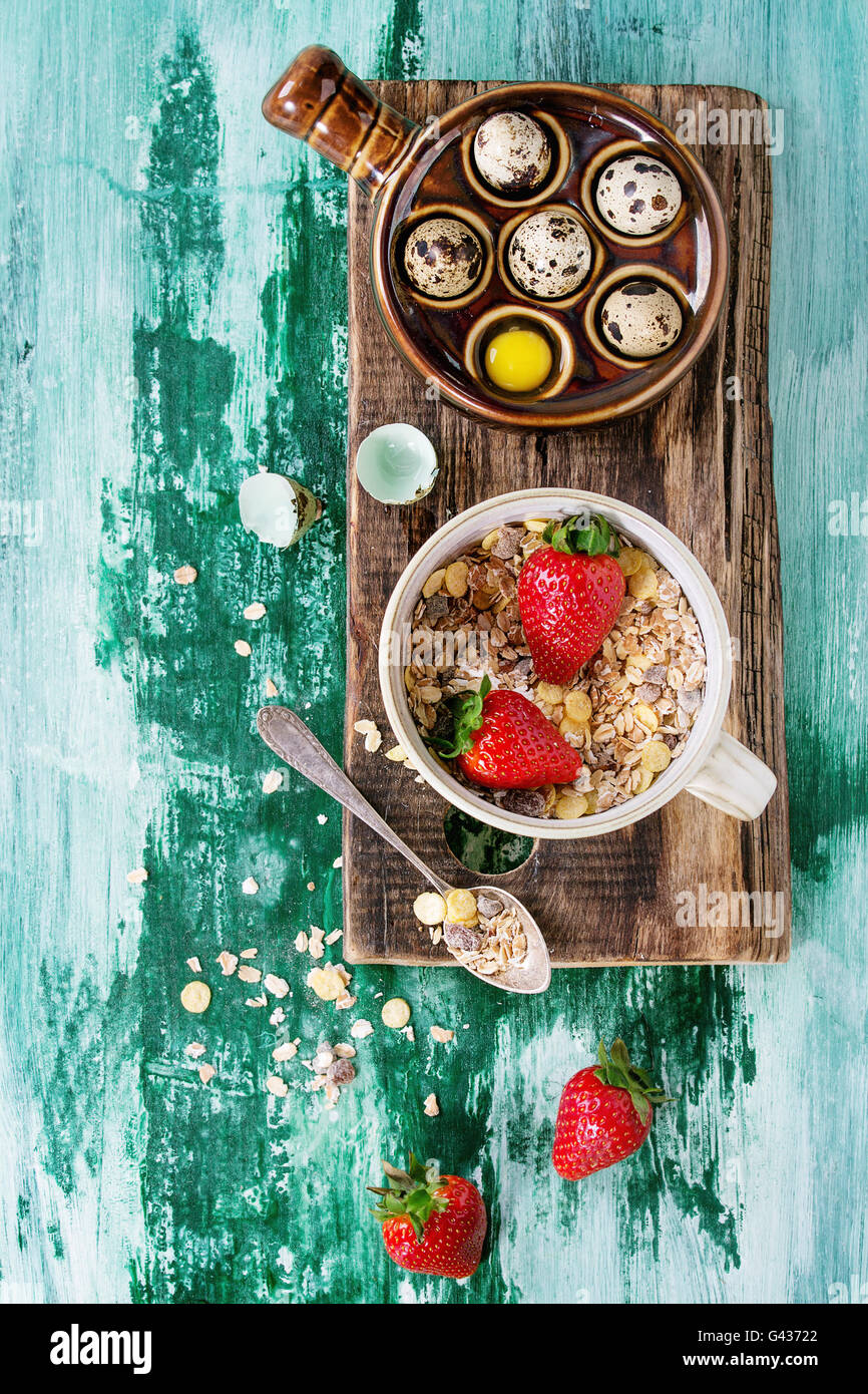 Ingredients for healthy breakfast. Quail eggs, bowl of muesli and strawberries on wood chopping board teaspoon over white and gr Stock Photo