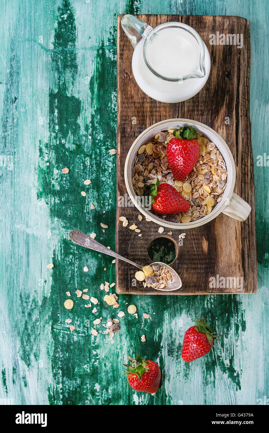Ingredients for healthy breakfast. Jug of milk, bowl of muesli and strawberries on wood chopping board teaspoon over white and g Stock Photo