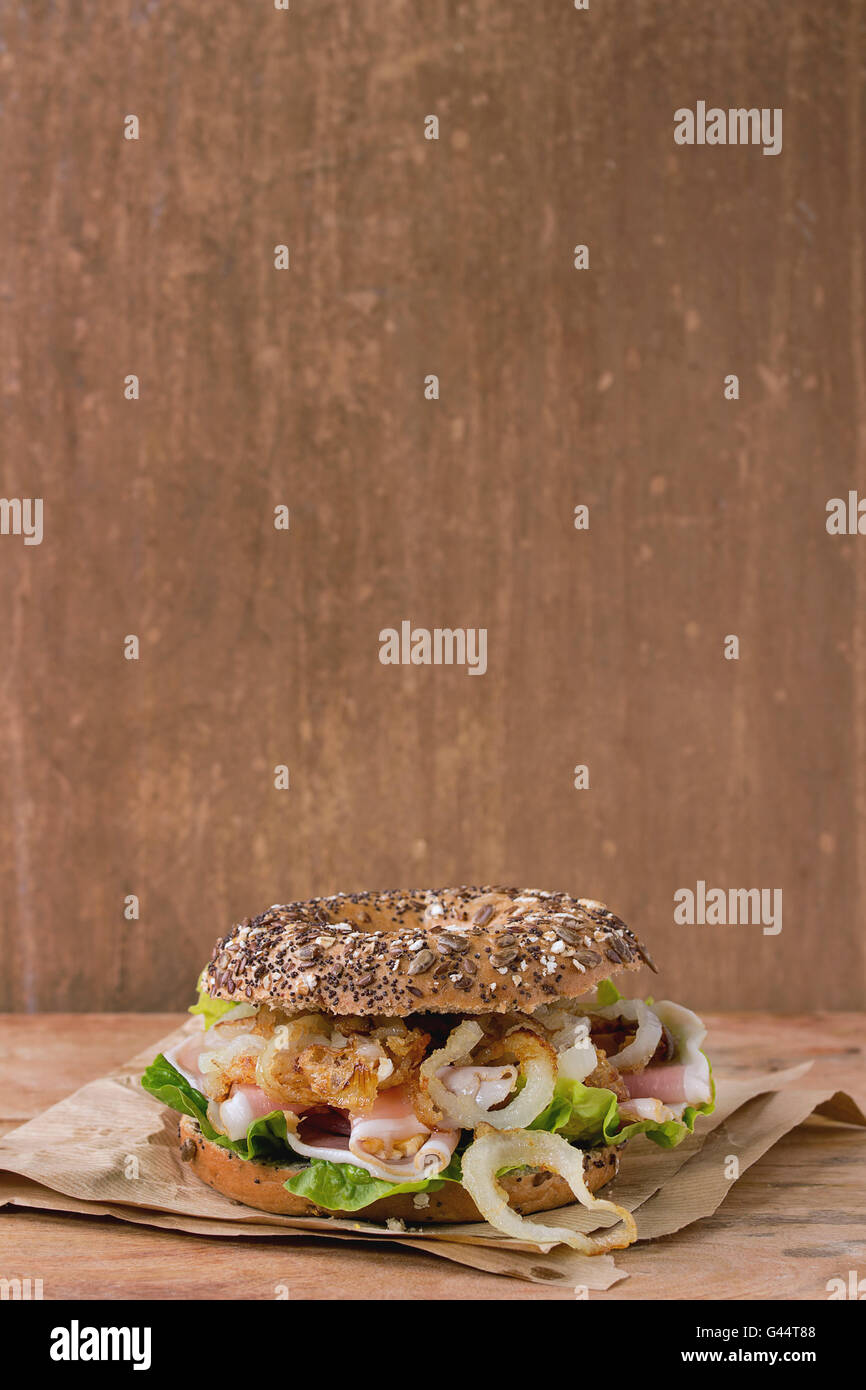 Whole Grain bagel with fried onion, green salad and prosciutto ham on paper over wooden textured background. With copy space Stock Photo