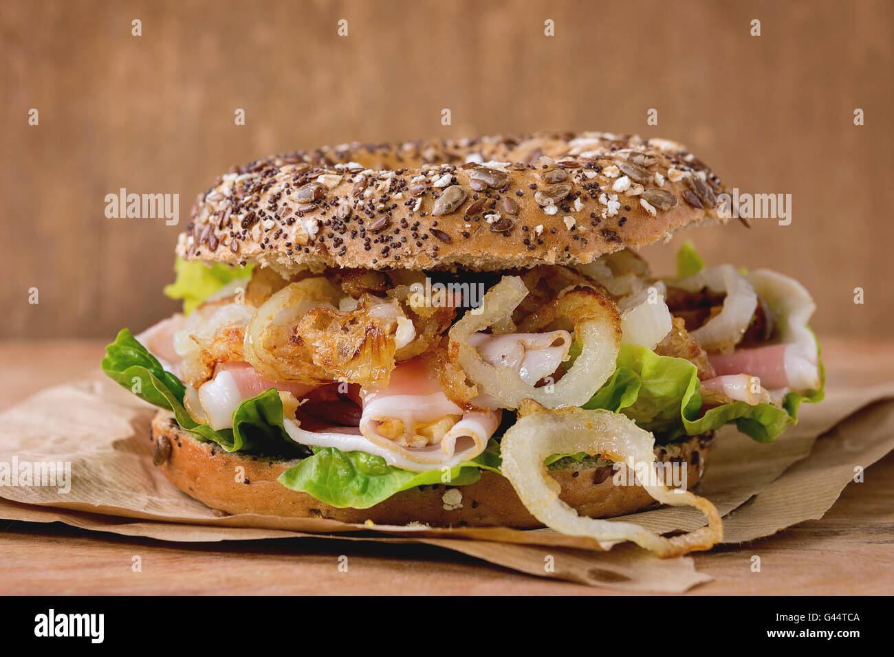 Whole Grain bagel with fried onion, green salad and prosciutto ham on paper over wooden textured background. Close up Stock Photo