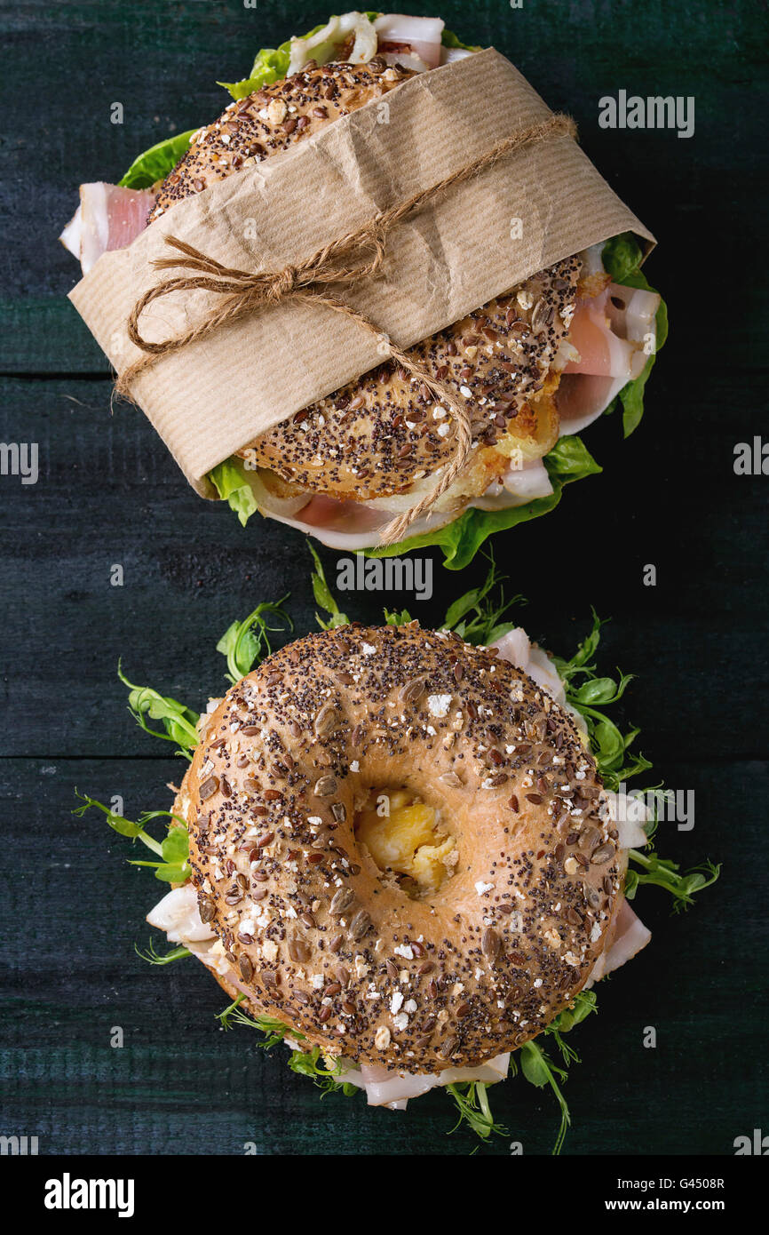Two Whole Grain bagels with fried onion, scrambled eggs, green salad and prosciutto ham over dark wooden textured background. To Stock Photo