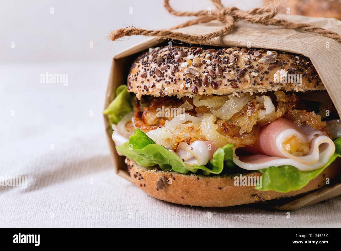 Papered Whole Grain bagel with fried onion, green salad and prosciutto ham over white linen tablecloth. Close up Stock Photo