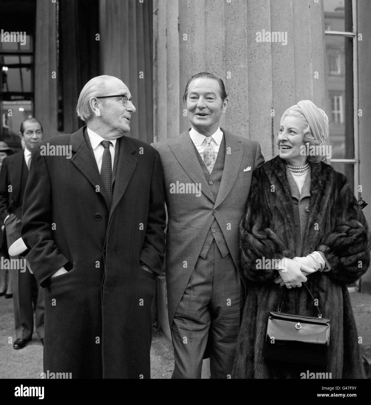 Playwright Sir Terence Rattigan (c) with producer Mr Harold French (l) and Mrs French (r) as he left Buckingham Palace after receiving the accolade as a Knight Bachelor from Queen Elizabeth the Queen Mother at an investiture. Mr French produced Mr Rattigan's first play. Stock Photo