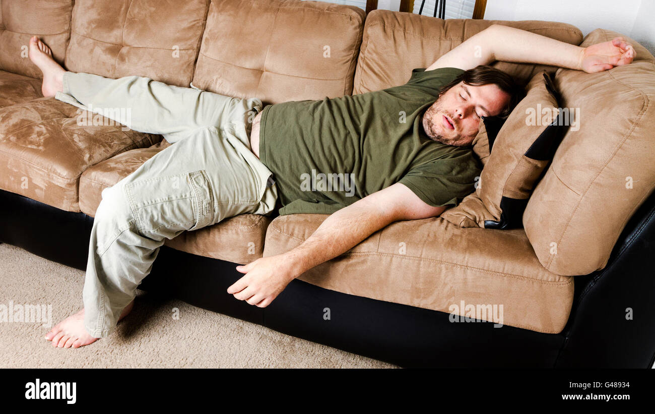 Single male sprawled out all over the couch sleeping with the TV on. Shows either relaxation or with the mans weight a lazy life Stock Photo