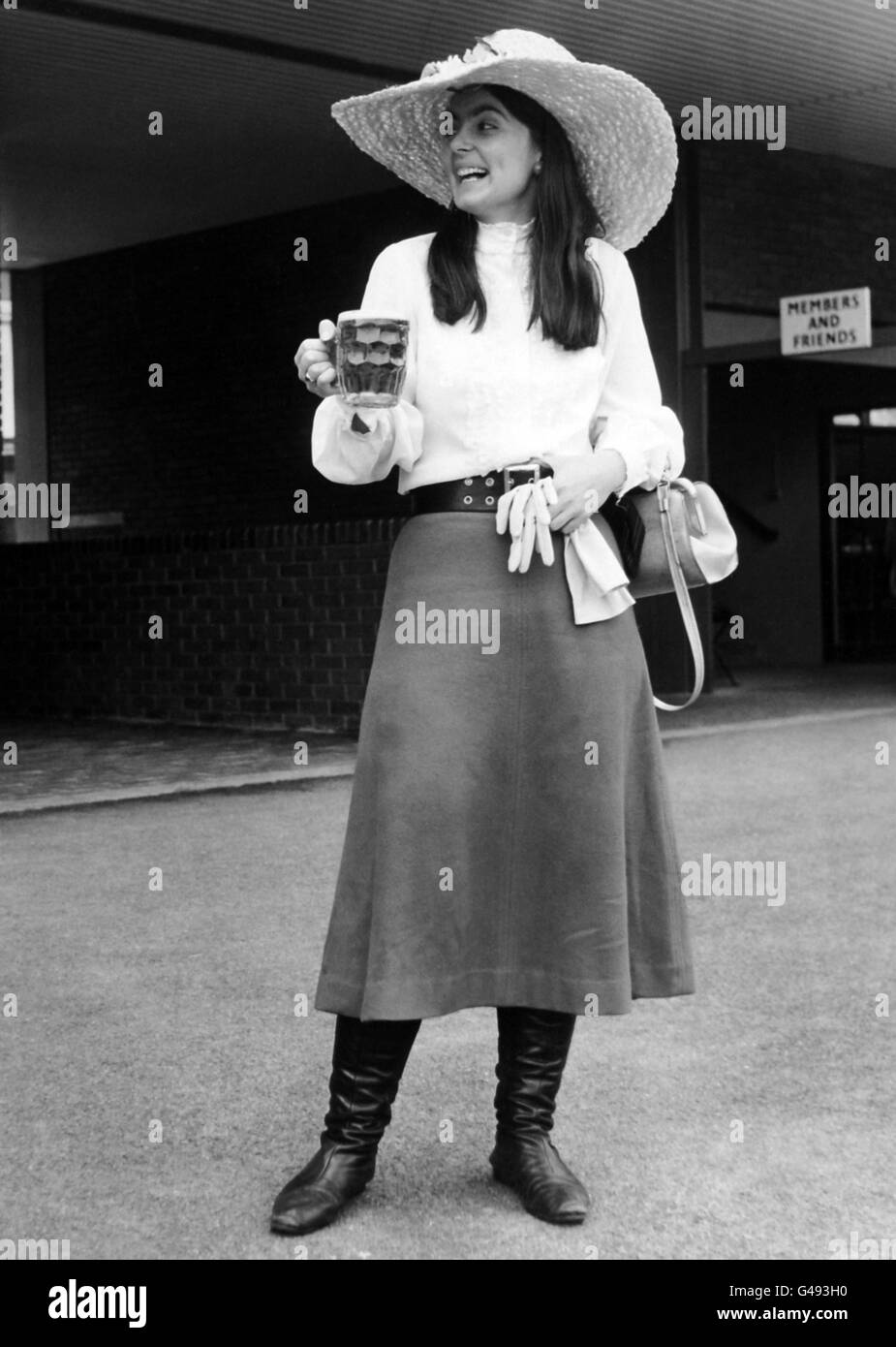 Nicky Webster from Harrow, whose brother plays for the Harrow team, wearing a maxi skirt and large straw hat trimmed with flowers. She drinks a pint of bitter while watching the match. Stock Photo