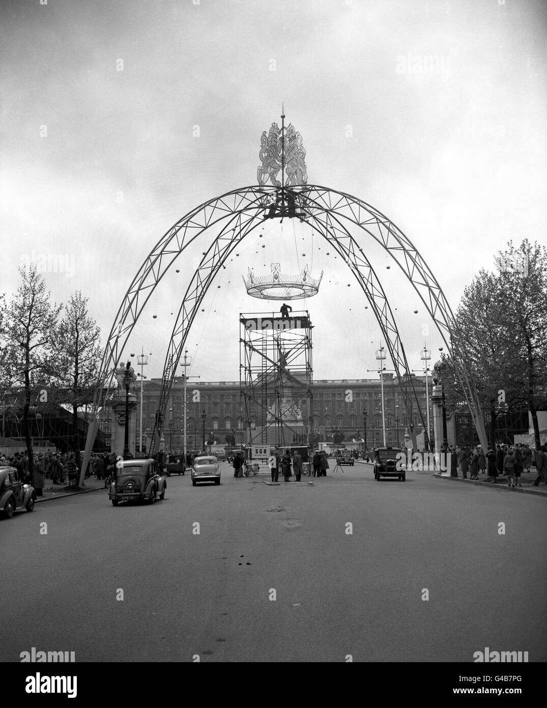 With crowds gathered on the pavements, and with Buckingham Palace as the backcloth, workmen hoist into position a replica of a princess's coronet, that will hang from a 65 foot arch - one of four to be part of the Coronation decorations in The Mall, London. Topping the arch are a lion and a unicorn, in steel tubing. Stock Photo