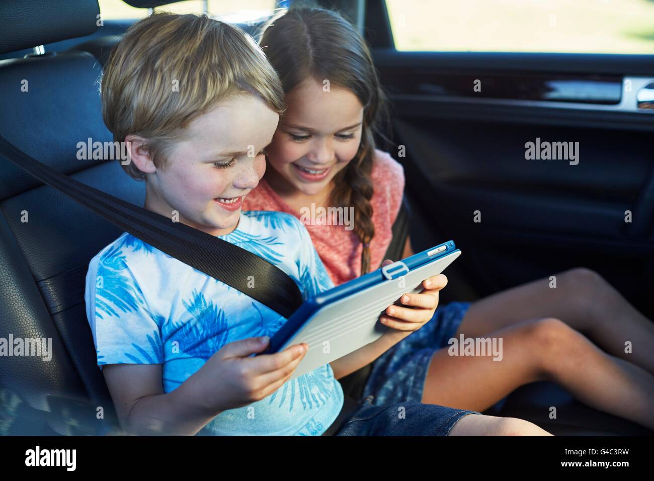 MODEL RELEASED. Brother and sister in the back seat of the car wearing seat belts, using a digital tablet. Stock Photo