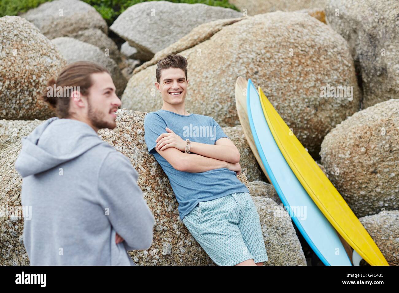 MODEL RELEASED. Young man leaning on rocks with surfboard. Stock Photo