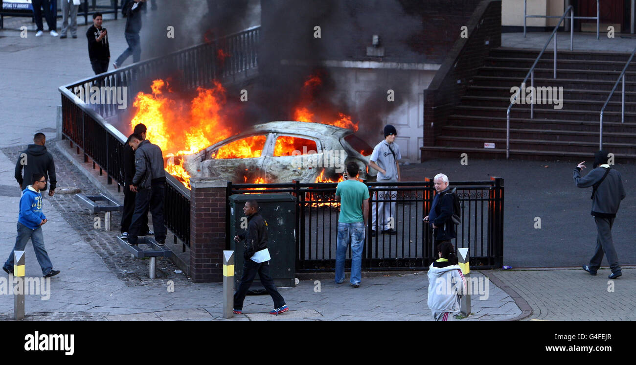 People pose for pictures near a burning car near Moor St Station and the Selfridges building in Birmingham, as fresh disturbances saw looting and vehicles set alight Birmingham. Stock Photo