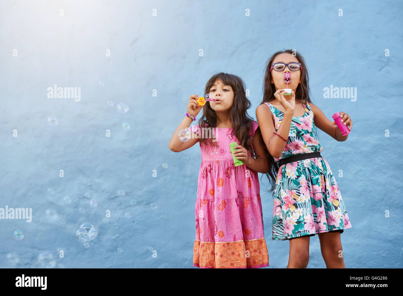 Portrait of adorable little girls blowing soap bubbles against blue wall. Two young girls playing together. Stock Photo