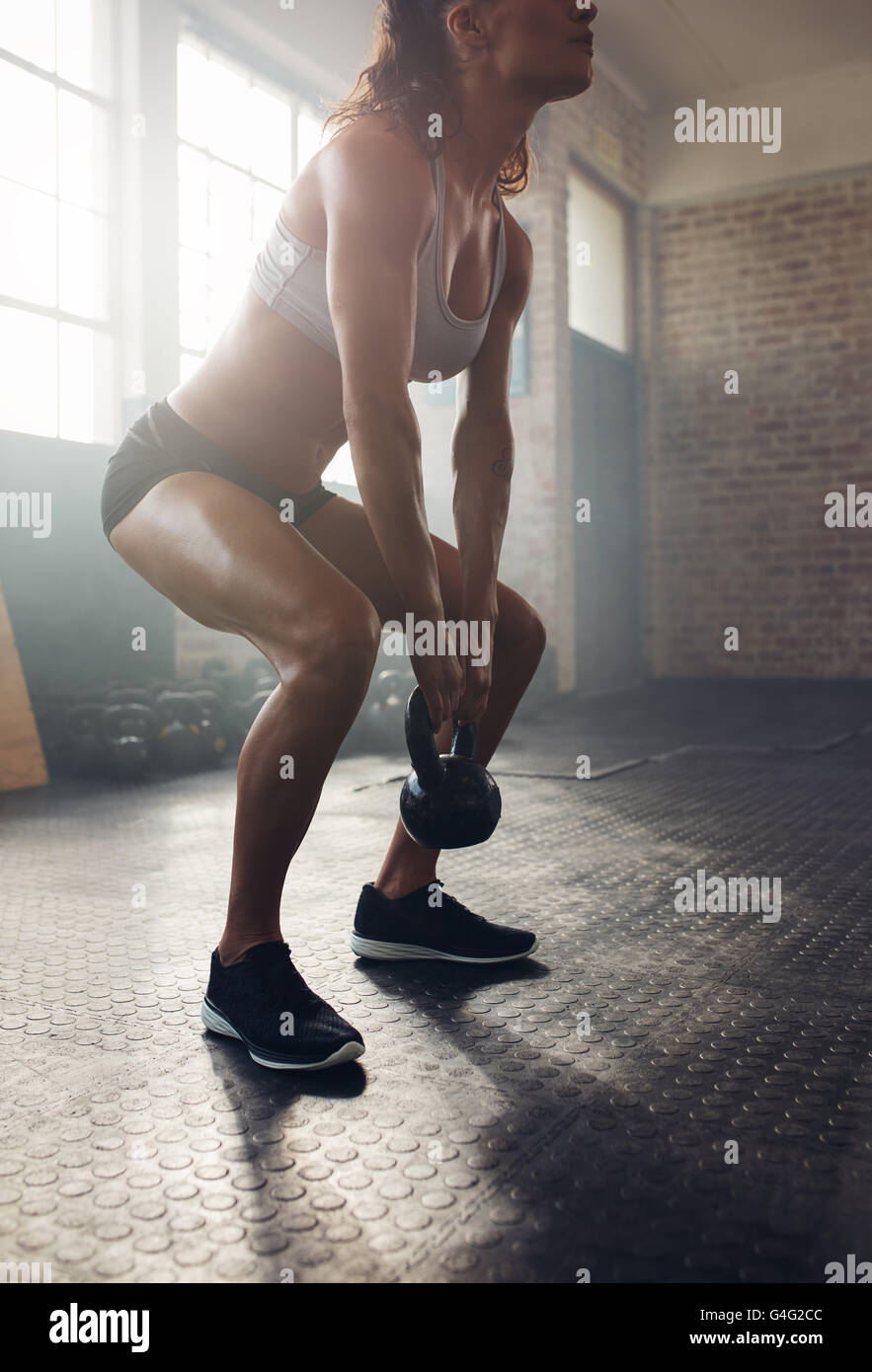 Athletic woman exercising with kettle bell while being in squat position. Muscular woman doing crossfit workout at gym. Stock Photo