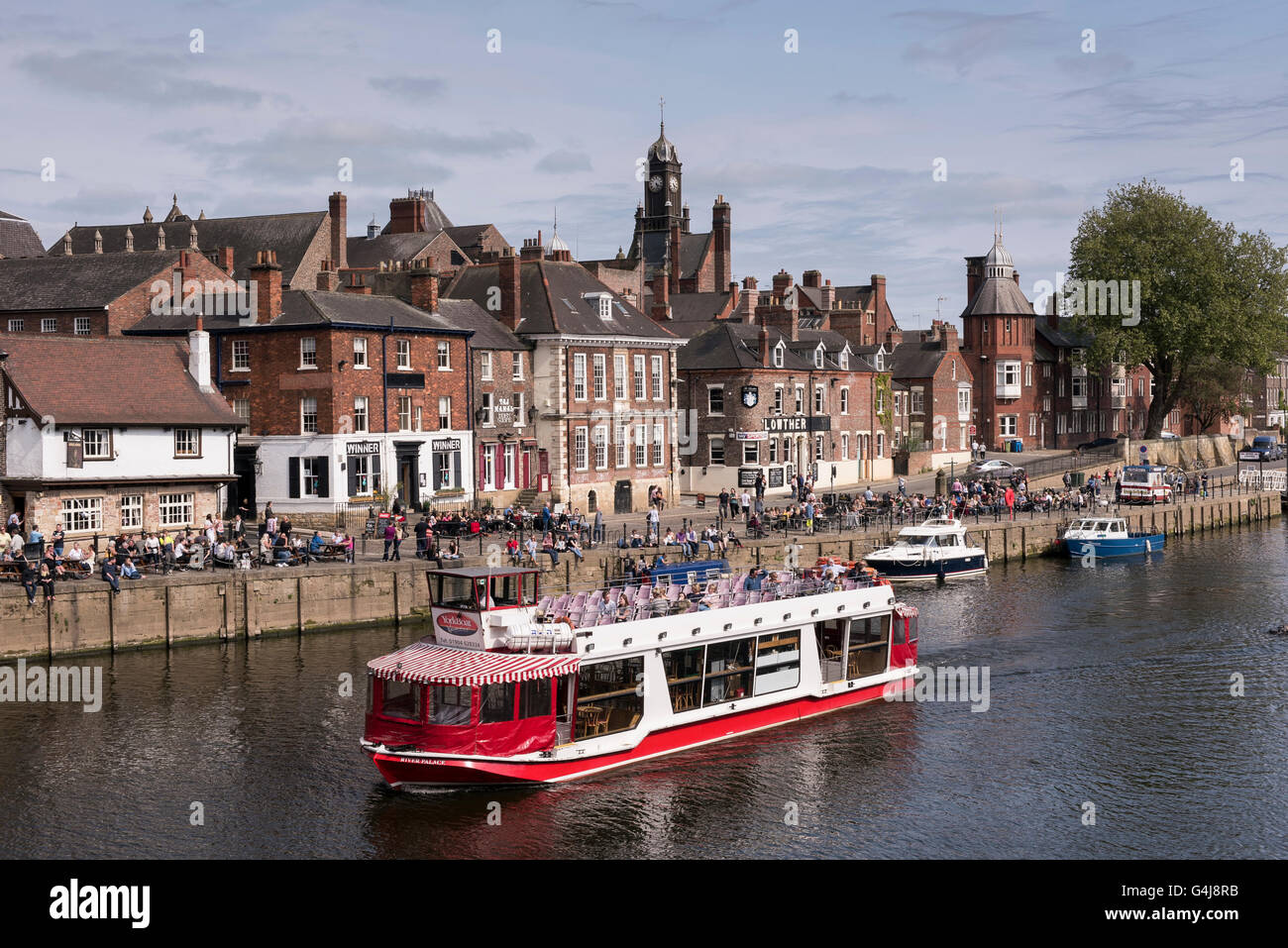 City Cruises boat sailing on sunny scenic River Ouse past people sitting relaxing outside riverside pubs & restaurants - York, Yorkshire, England, UK. Stock Photo