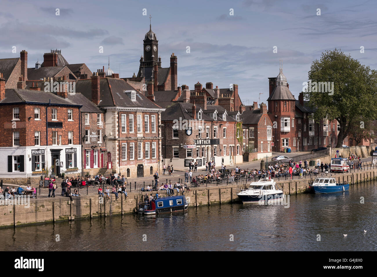 Lots of people drinking & relaxing in sun at busy riverside pubs & leisure boats moored on River Ouse - King's Staith, York, North Yorkshire, England. Stock Photo