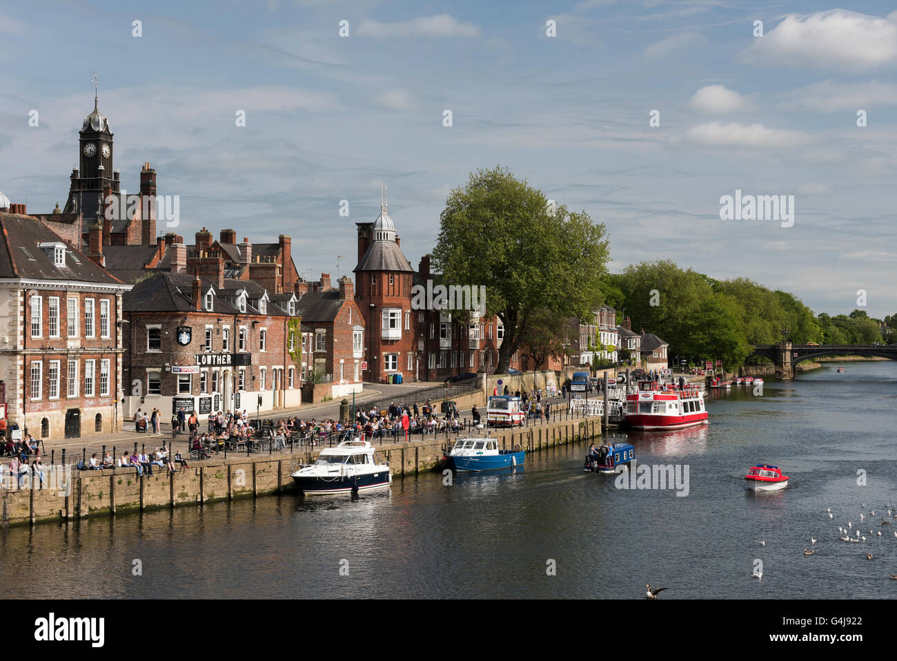 Lots of people drinking & relaxing in sun at busy riverside pub as leisure boats travel on River Ouse - King's Staith, York, North Yorkshire, England. Stock Photo