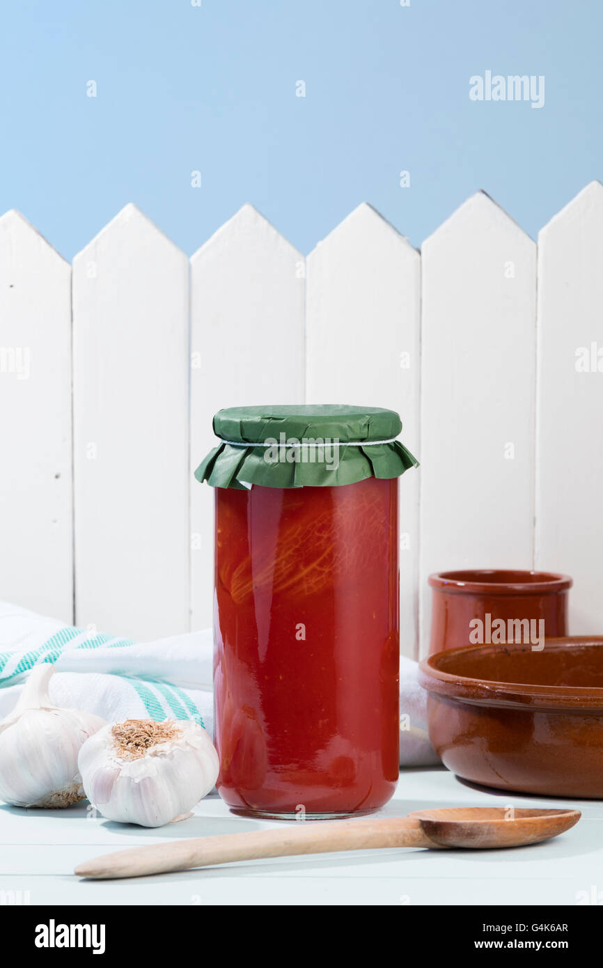 tomato jar on a wooden table with some some kitchen objects like a wooden spoon, dishes, garlics and a white napkin Stock Photo