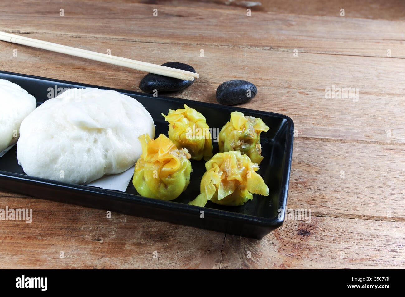 Buns of Pork stuffed and dumplings on a ceramic black plate. Bamboo chopsticks placed on a black stone. Stock Photo