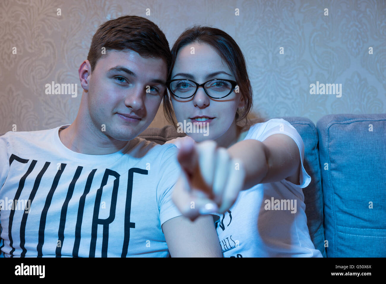 Attractive young girl is pointing at the camera sitting on a sofa with her boyfriend at home watching television, frontal view in the blue glow from the set. Stock Photo