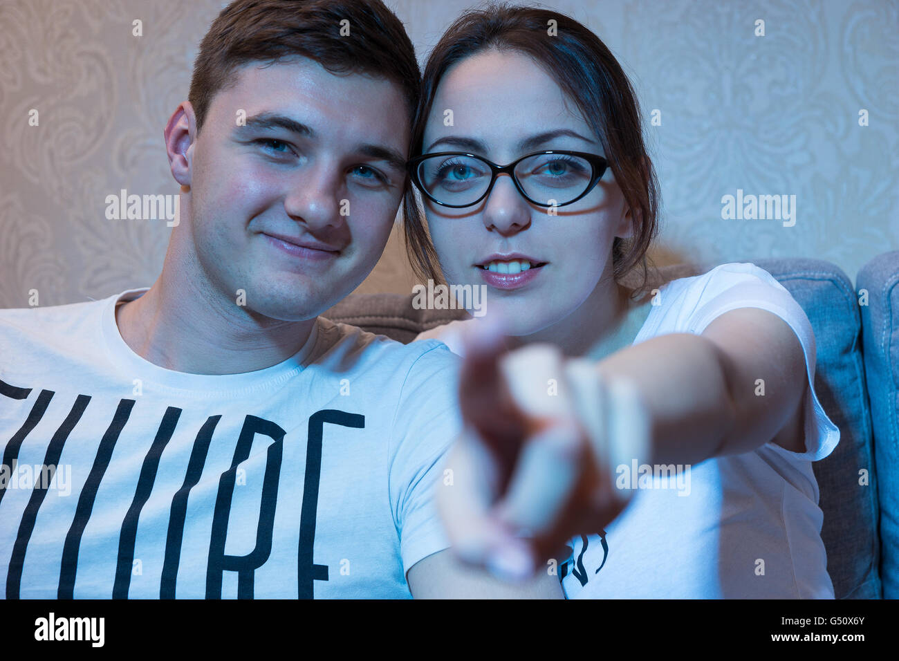 Young beautiful girl in glasses is pointing at the camera sitting on a sofa with her boyfriend at home watching television, frontal view in the blue glow from the set. Stock Photo