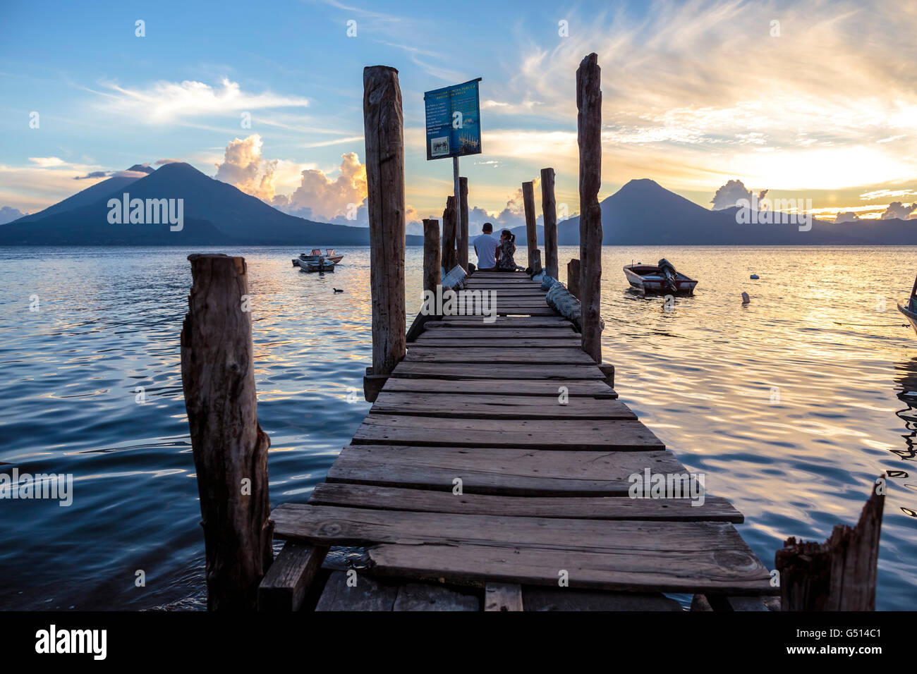 A couple of tourists sit on the dock in Panajachel as the sun sets over Lago de Atitlan, a volcanic crater lake in Guatemala Stock Photo