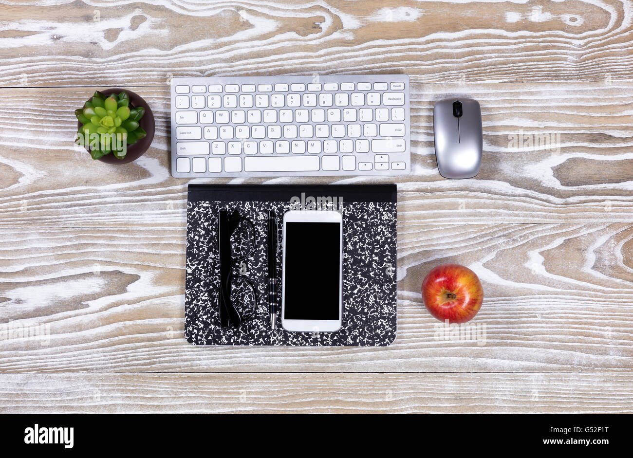 Old desktop with notepad, pen, apple, computer keyboard, plant, mouse, reading glasses, apple and cell phone. Stock Photo