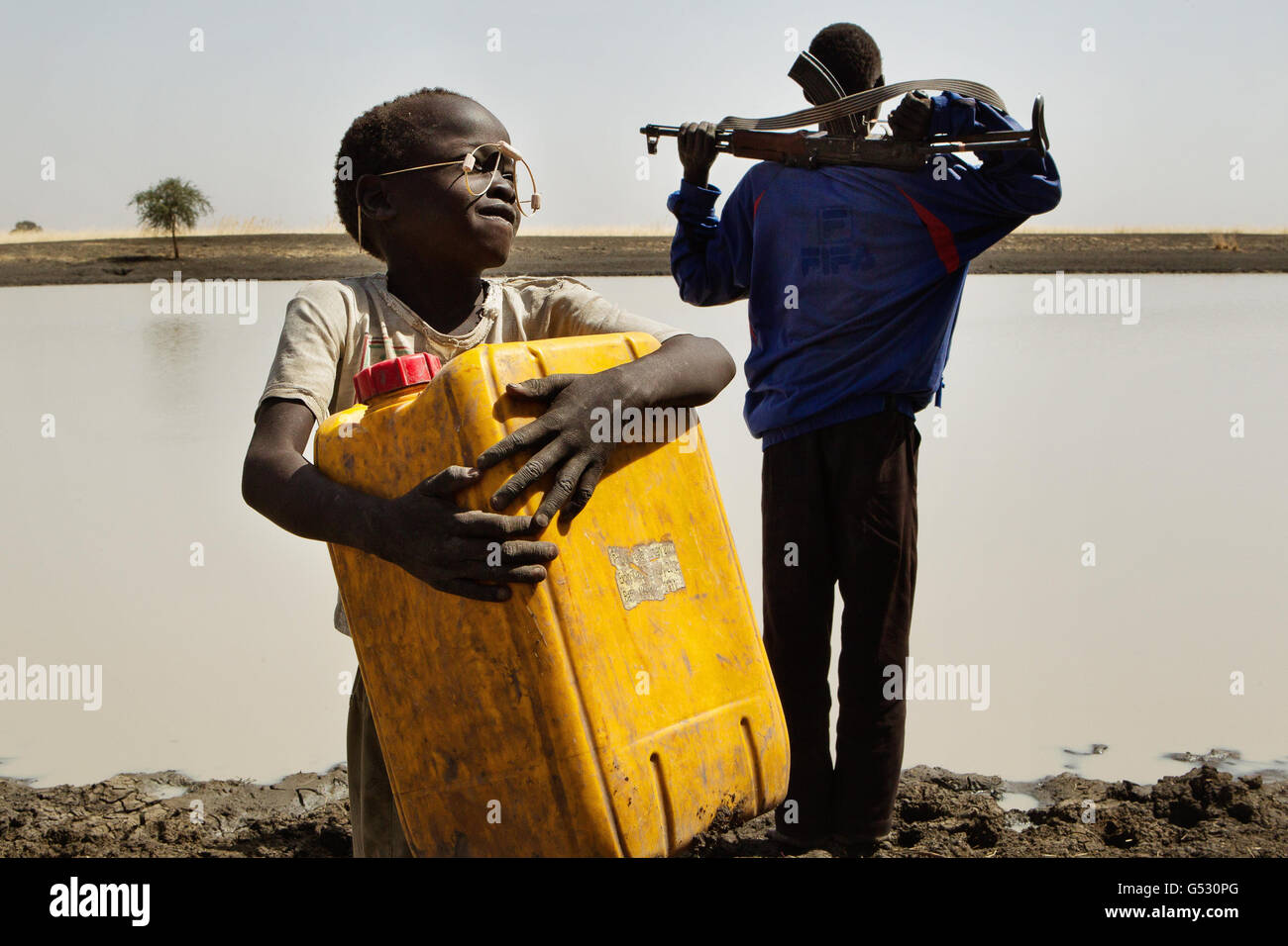 A young boy carries water in a jerry can to his village while his grandfather, the village chief, cradles an AK 47 rifle watching out for cattle rustlers and lions, at the only water source in the area, along the Sobat River in the Greater Upper Nile region of north-eastern South Sudan, Africa. Stock Photo