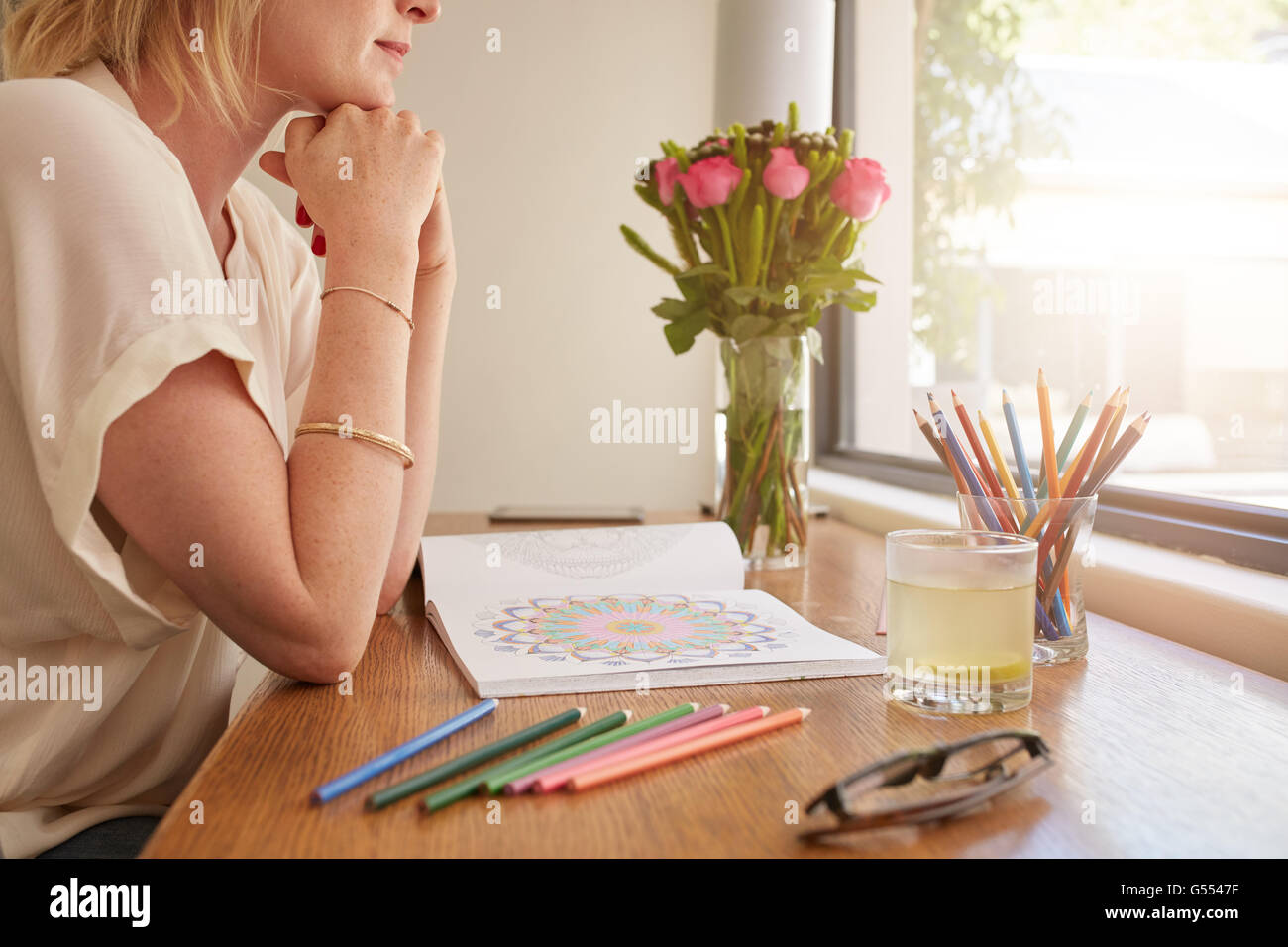 Side view cropped shot of young woman sitting at a table and thinking with adult coloring book and pencils colors. Stock Photo